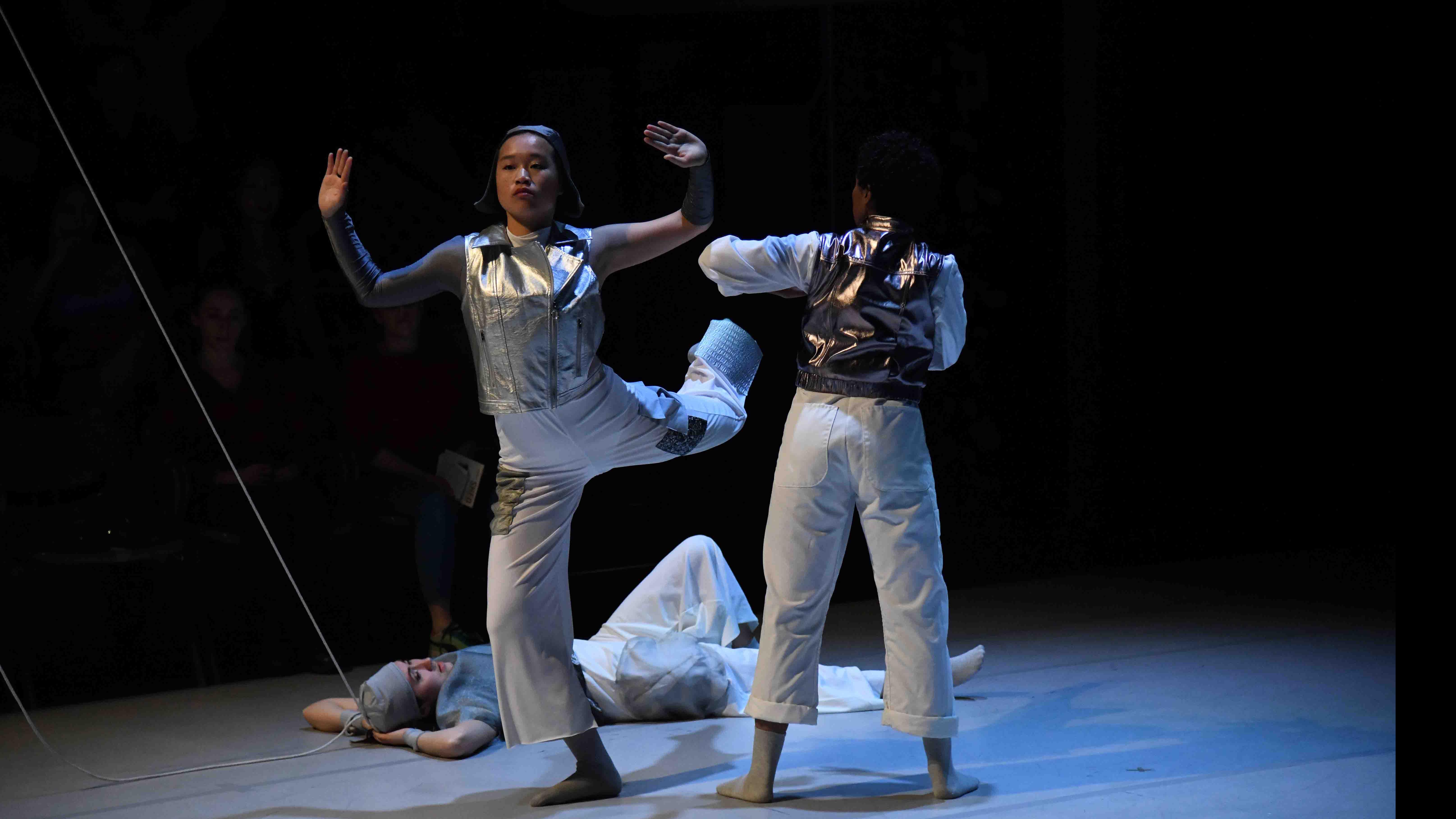 Two student dancers in movement poses, one facing the camera, one facing away, and a third student dancer lying on the ground, with ropes looping down onto the stage. Photo credit: Thomas Hoebbel