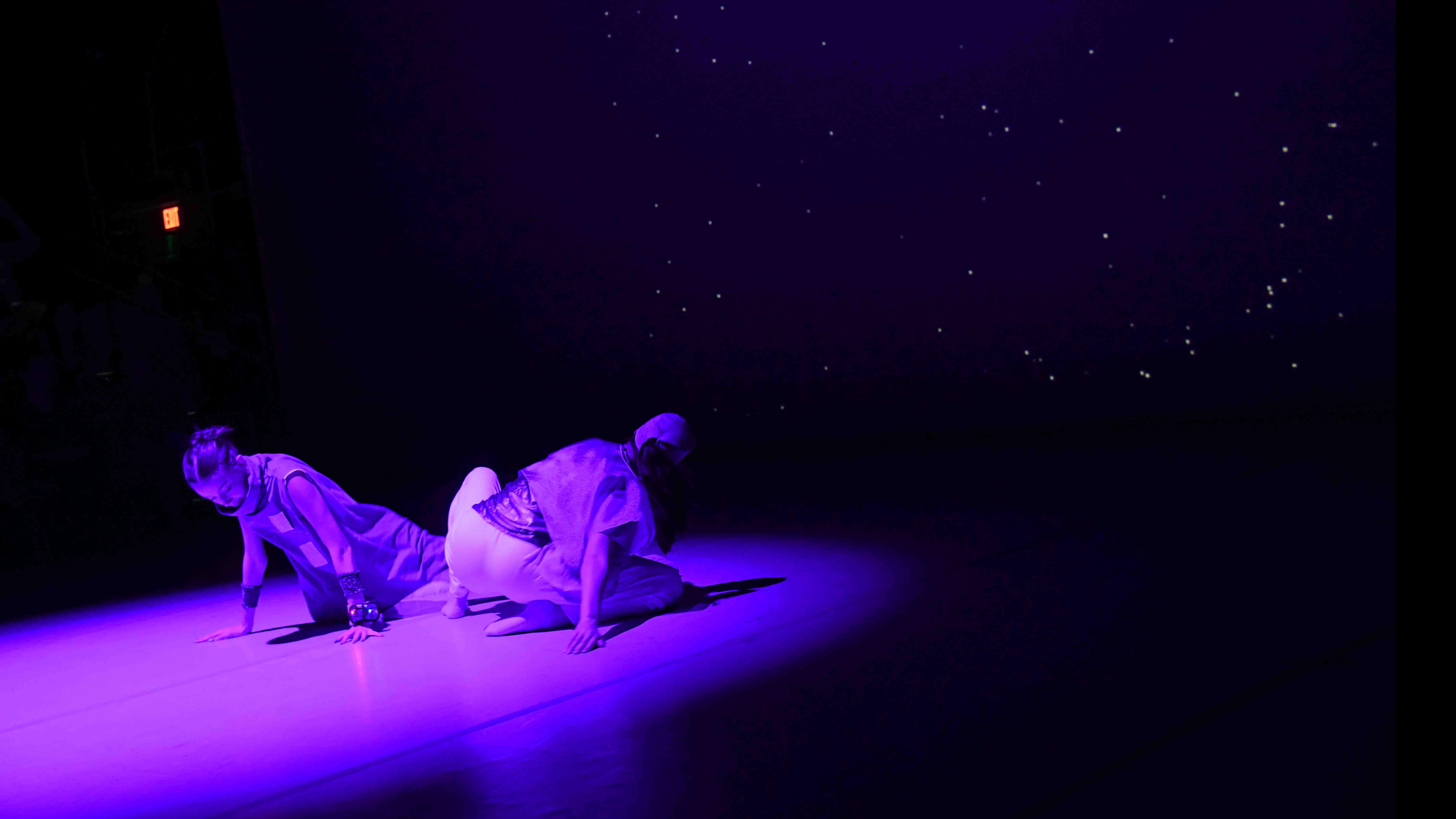 Two student dancers crouched face away from each other in a purple spotlight on stage. Photo credit: Thomas Hoebbel
