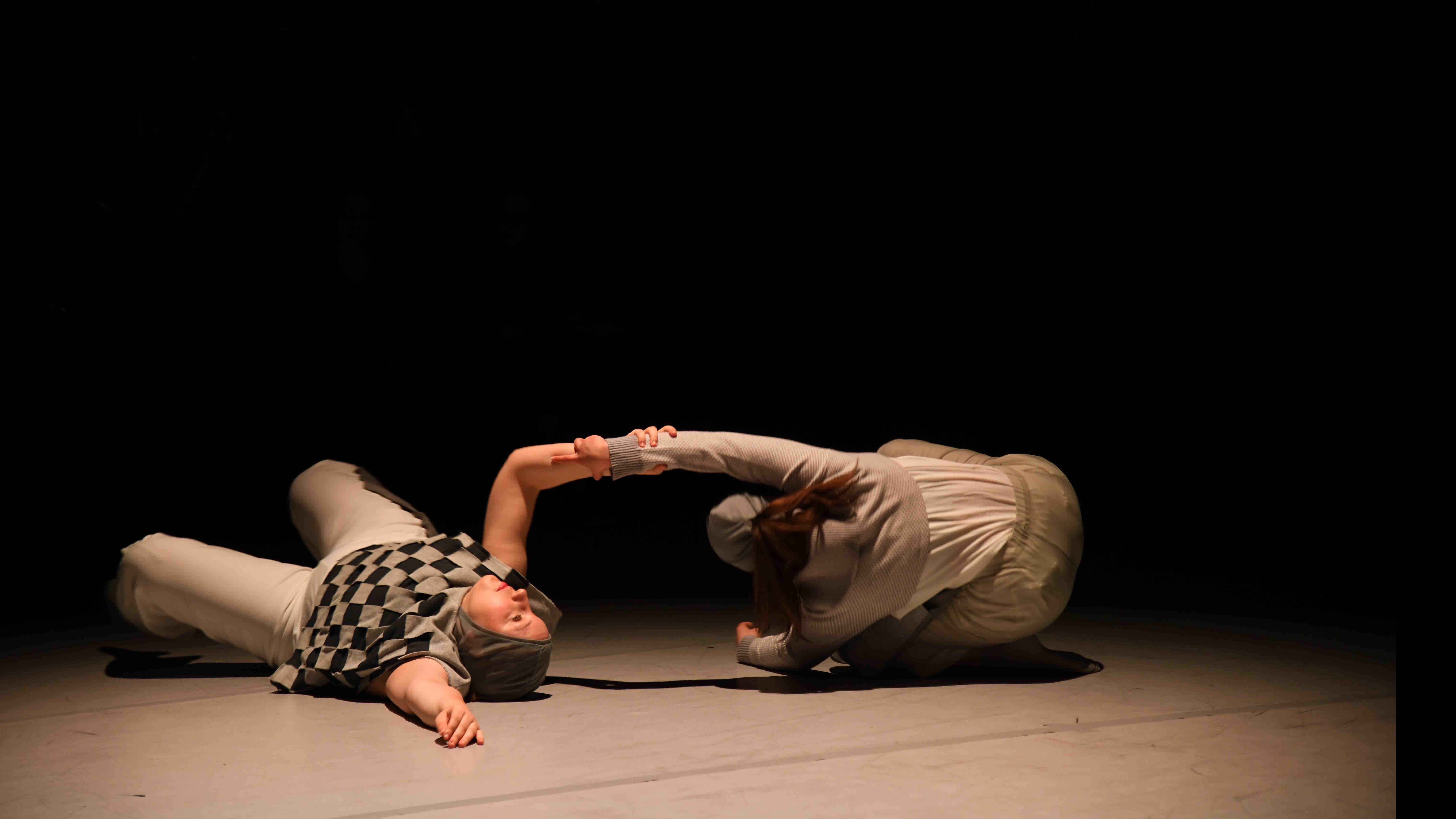 Two student dancers in movement poses on the ground interlock arms in a spotlight on stage. Photo credit: Thomas Hoebbel