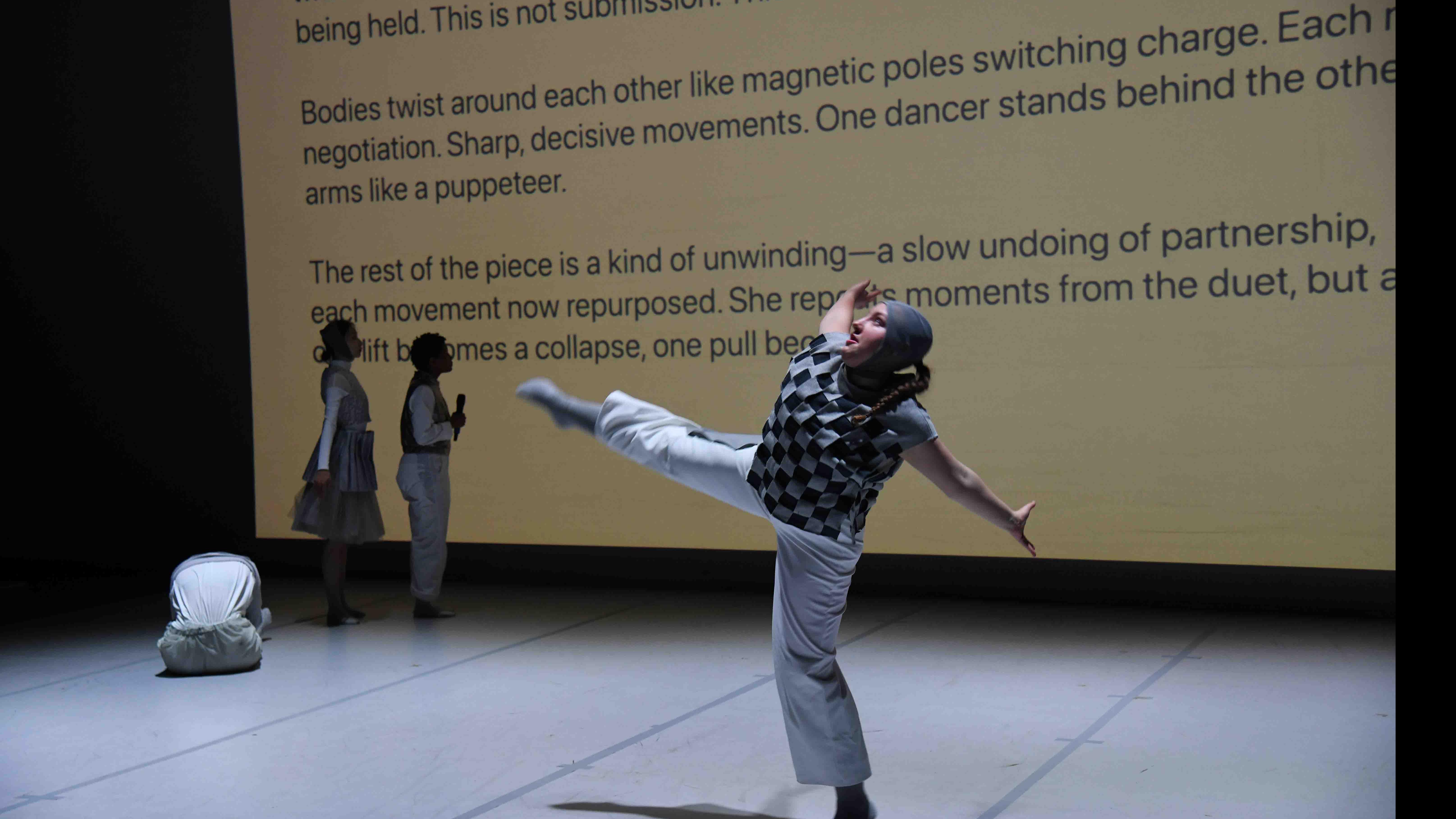 In the foreground, a student dancer extends their arms and a leg in a movement pose, and in the background three student dancers stand and look at a screen showing these words: “…twitch. There’s unrest beneath the…being held. This is not submission. This is burden. From here, the…. Bodies twist around each other like magnetic poles switching charge. Each… negotiation. Sharp, decisive movements. One dancer stands behind the other… arms like a puppeteer..." Photo credit: Thomas Hoebbel