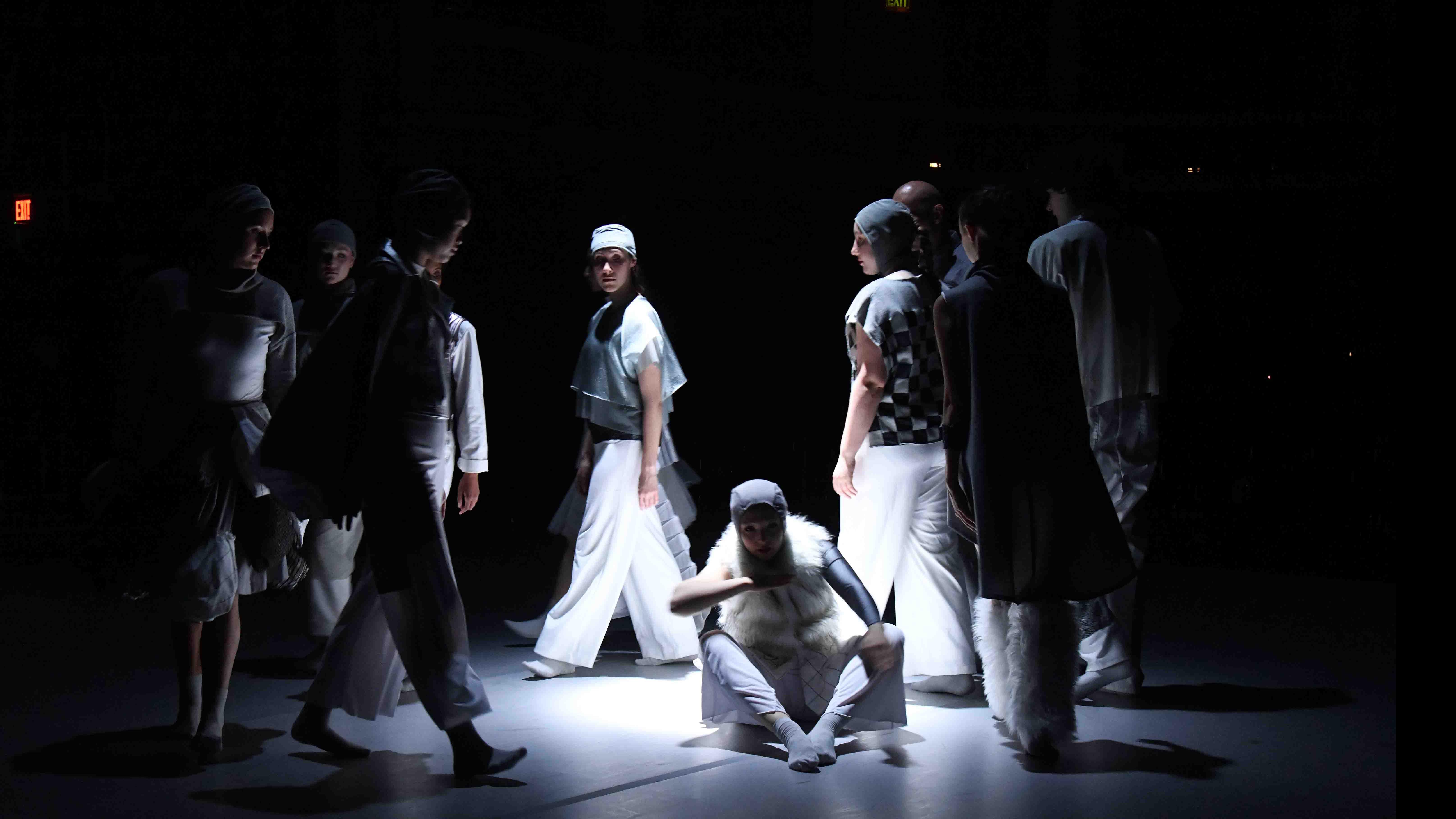 An ensemble of student dancers encircles a student dancer who sits on the ground on stage in a spotlight. Photo credit: Thomas Hoebbel