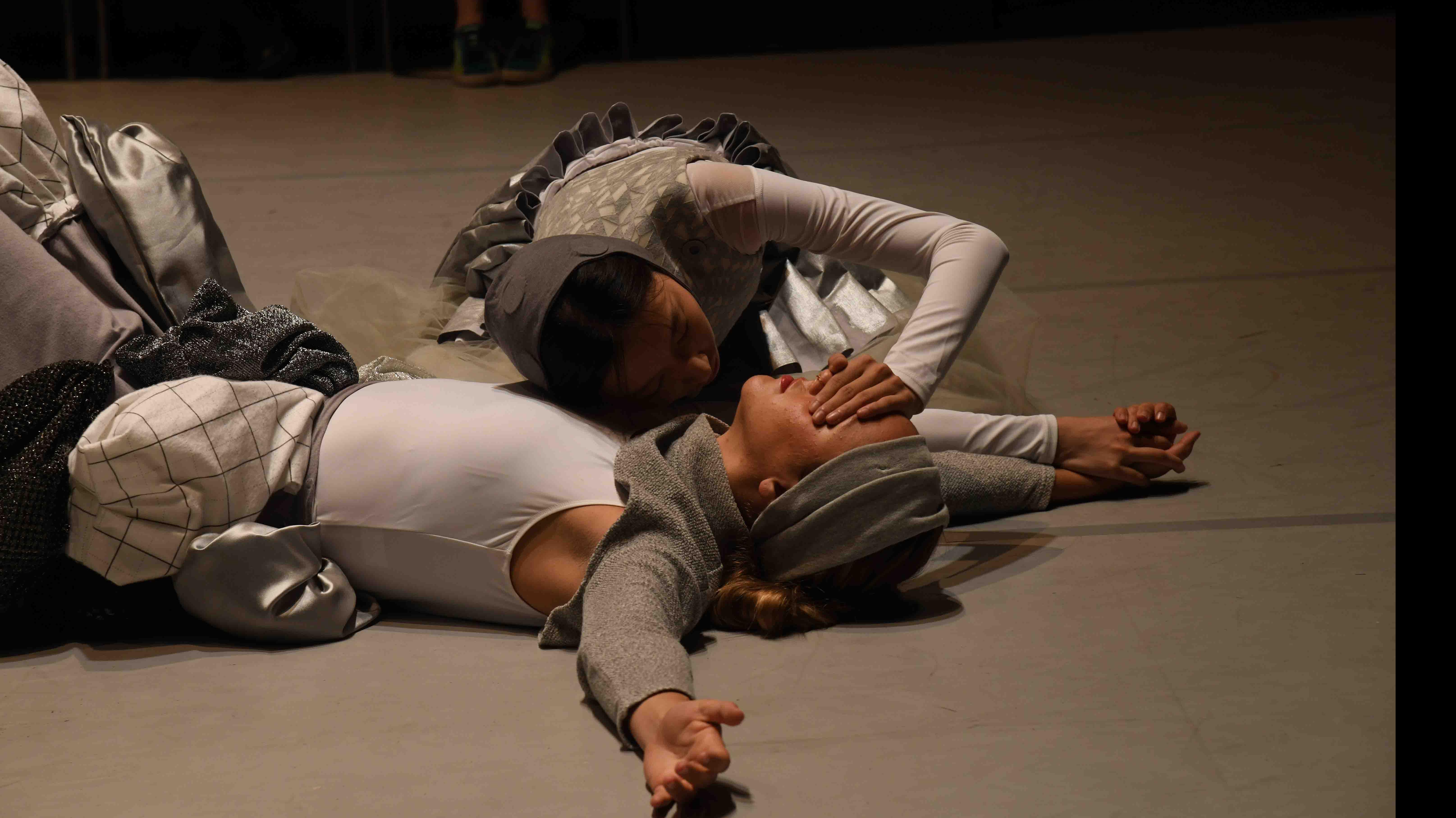Two student dancers on the ground on stage, one with arms outstretched, and one covering the eyes of the first. Photo credit: Thomas Hoebbel