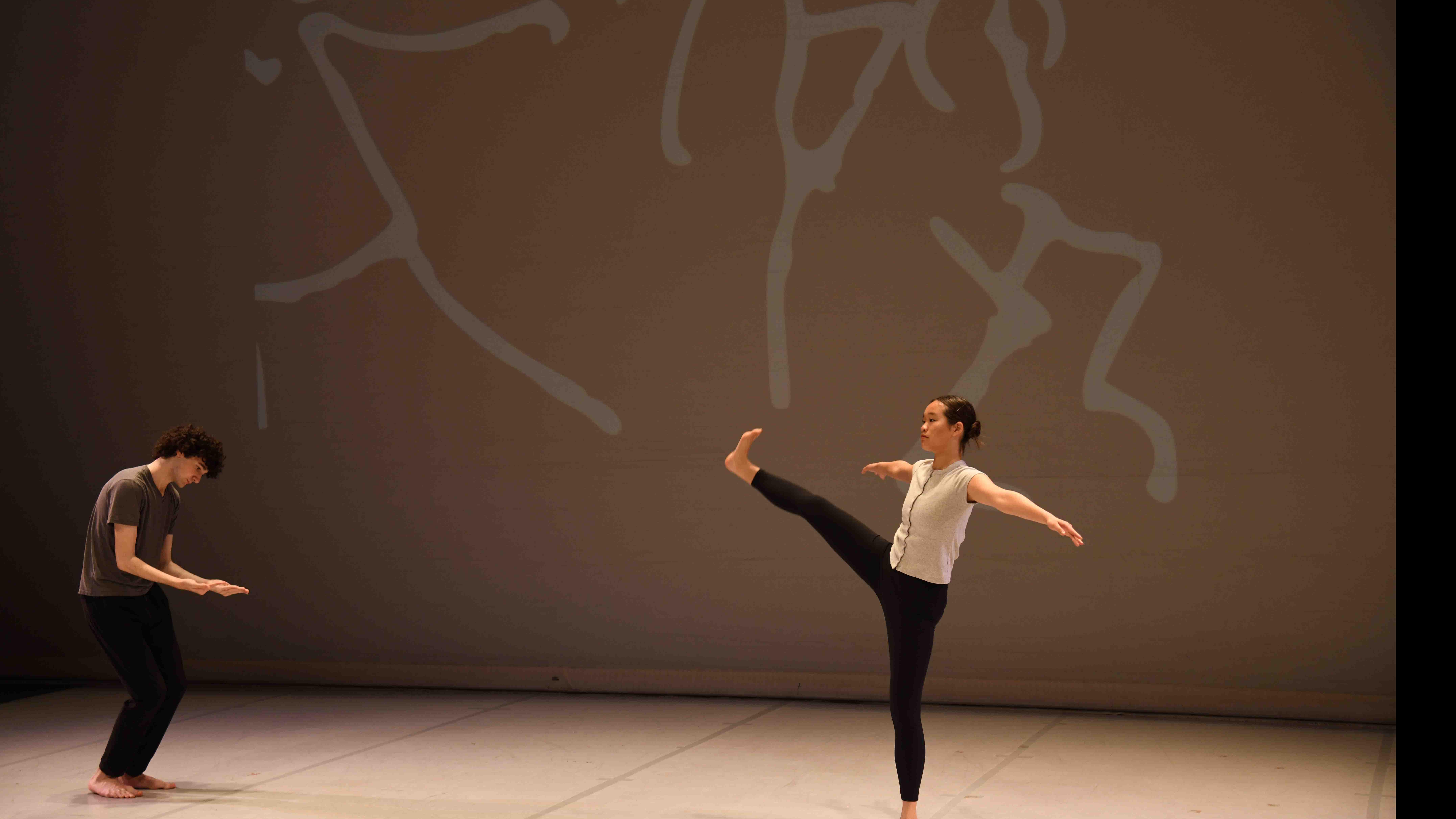 On left, a student dancer crouches, and on right, a student dancer kicks out a leg and arms, with hand drawn figures on a screen behind them, on stage. Photo credit: Thomas Hoebbel