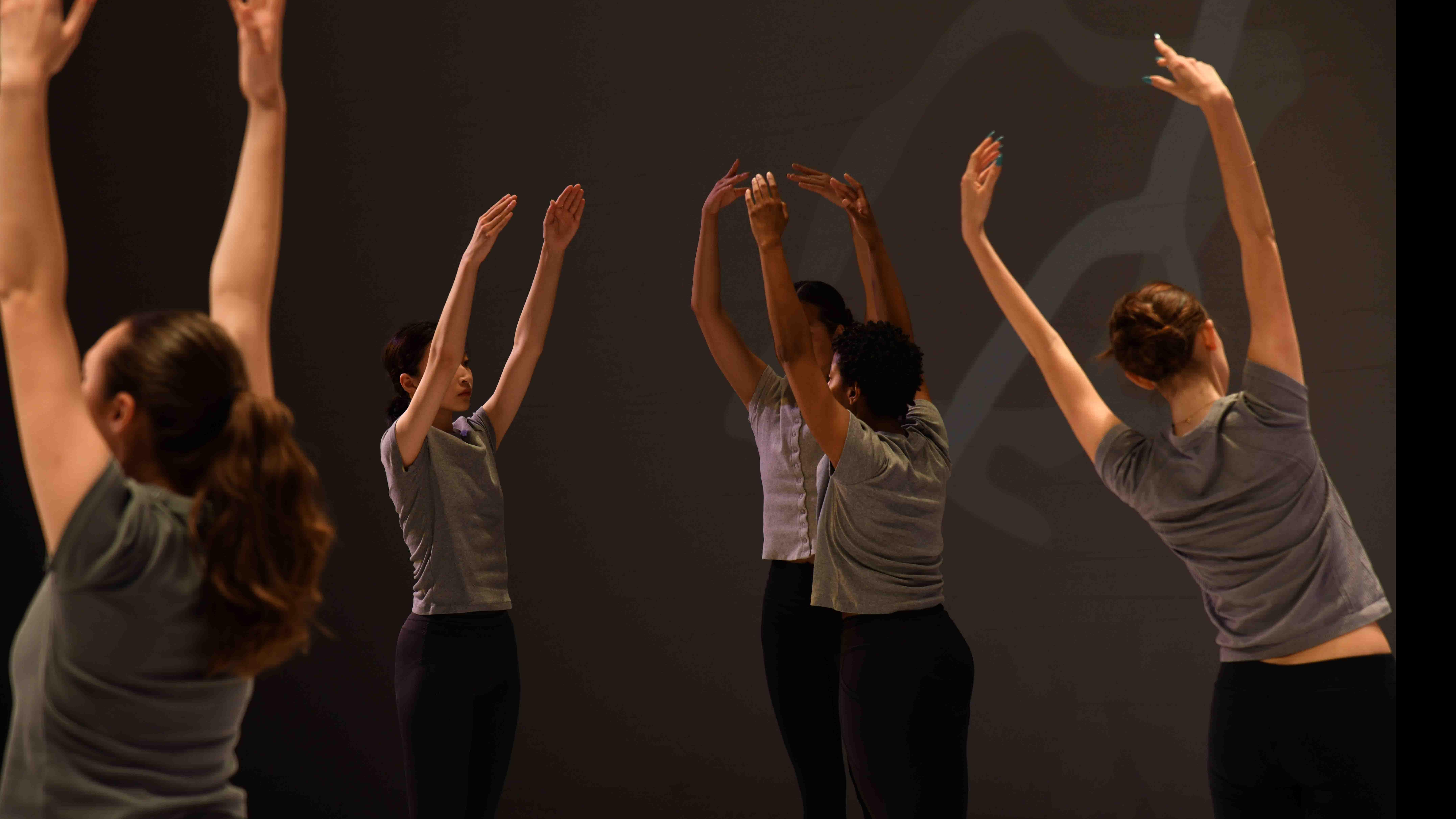 Five student dancers facing different directions reach their arms up in identical movement poses, on stage. Photo credit: Thomas Hoebbel
