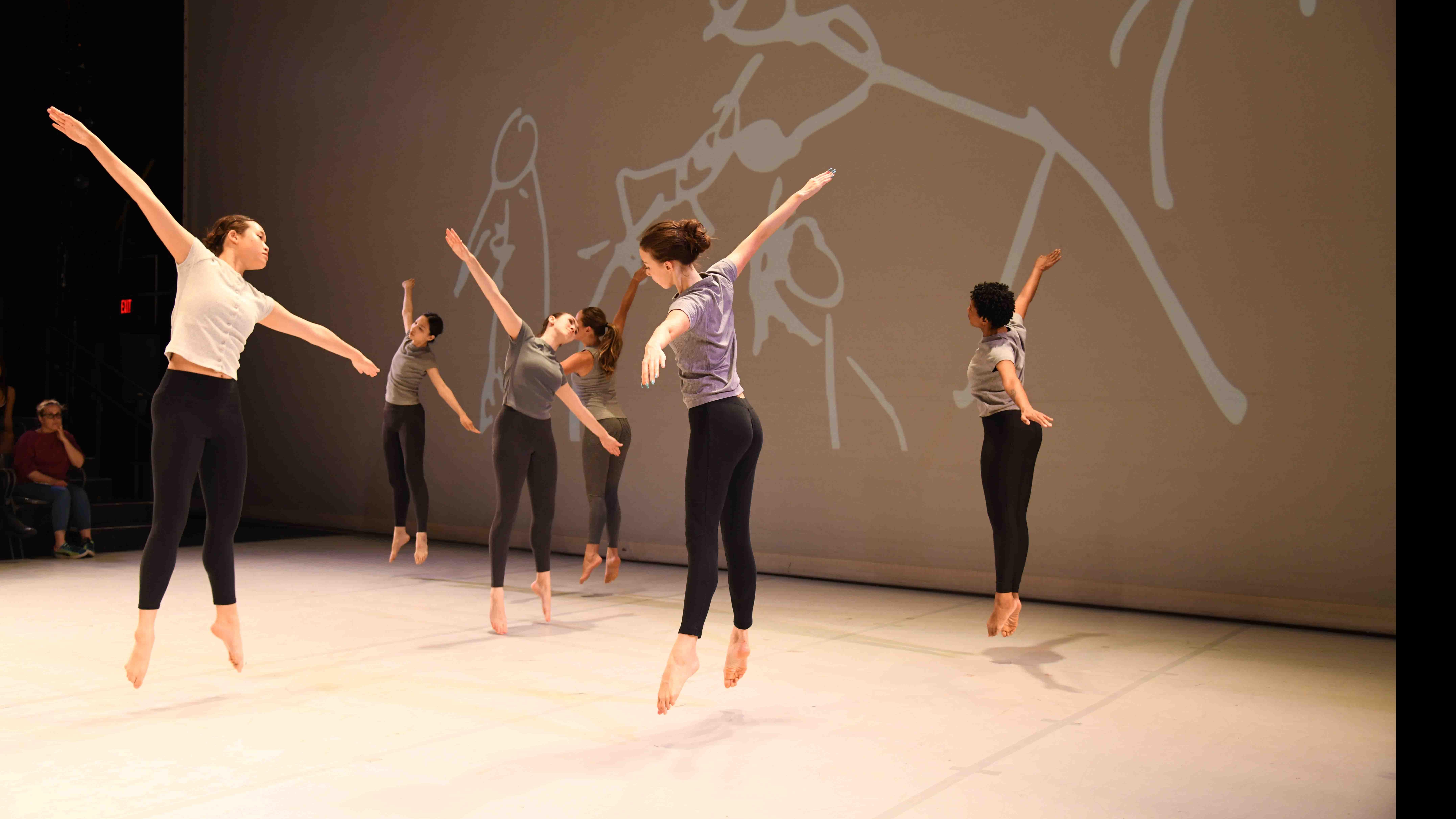 Six student dancers facing different directions jump in the air with arms diagonally outstretched in identical movement poses, with hand drawn figures on a screen behind them on stage. Photo credit: Thomas Hoebbel