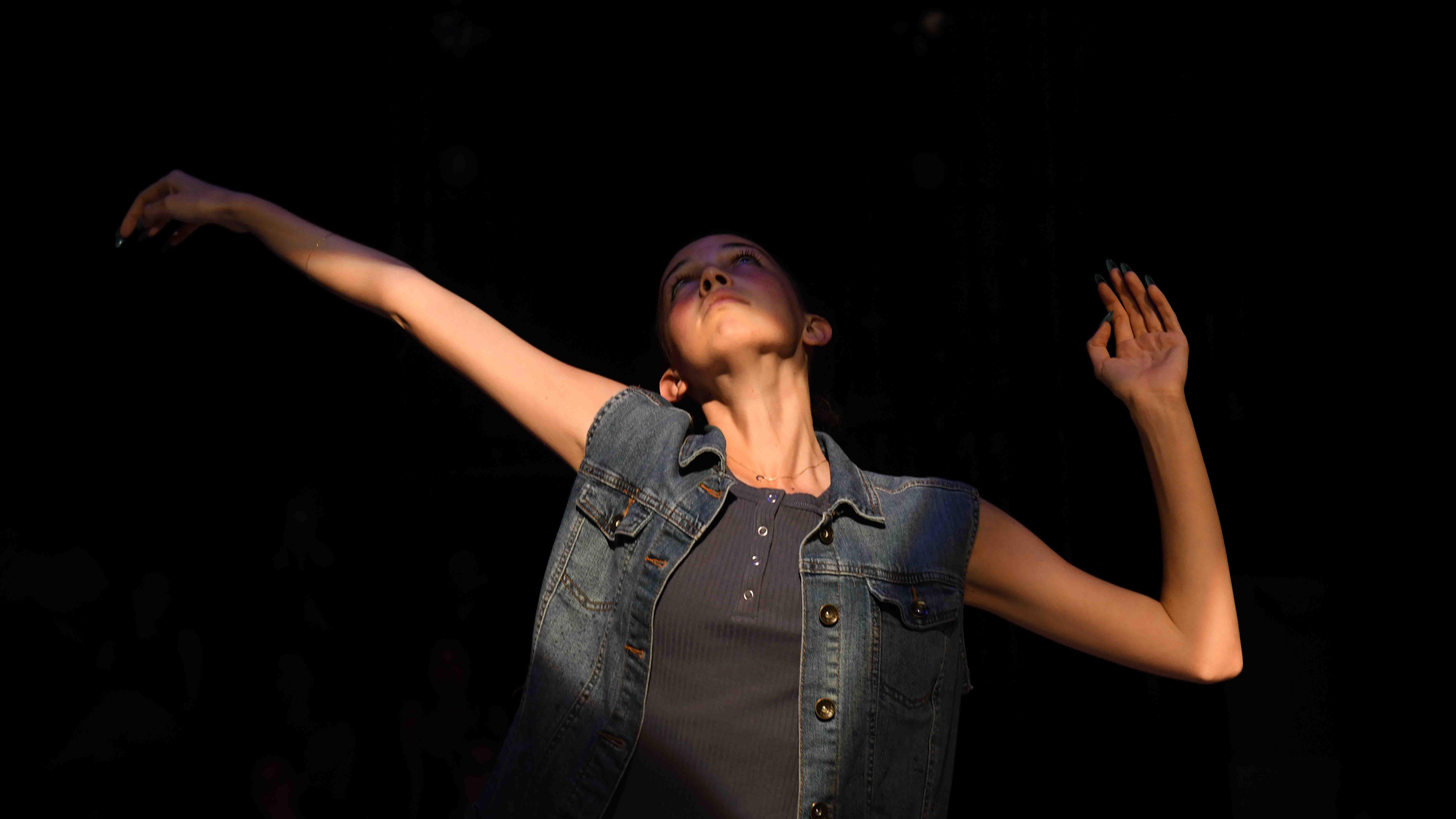 A student dancer stands with one arm bent and one arm outstretched looking up on stage. Photo credit: Thomas Hoebbel