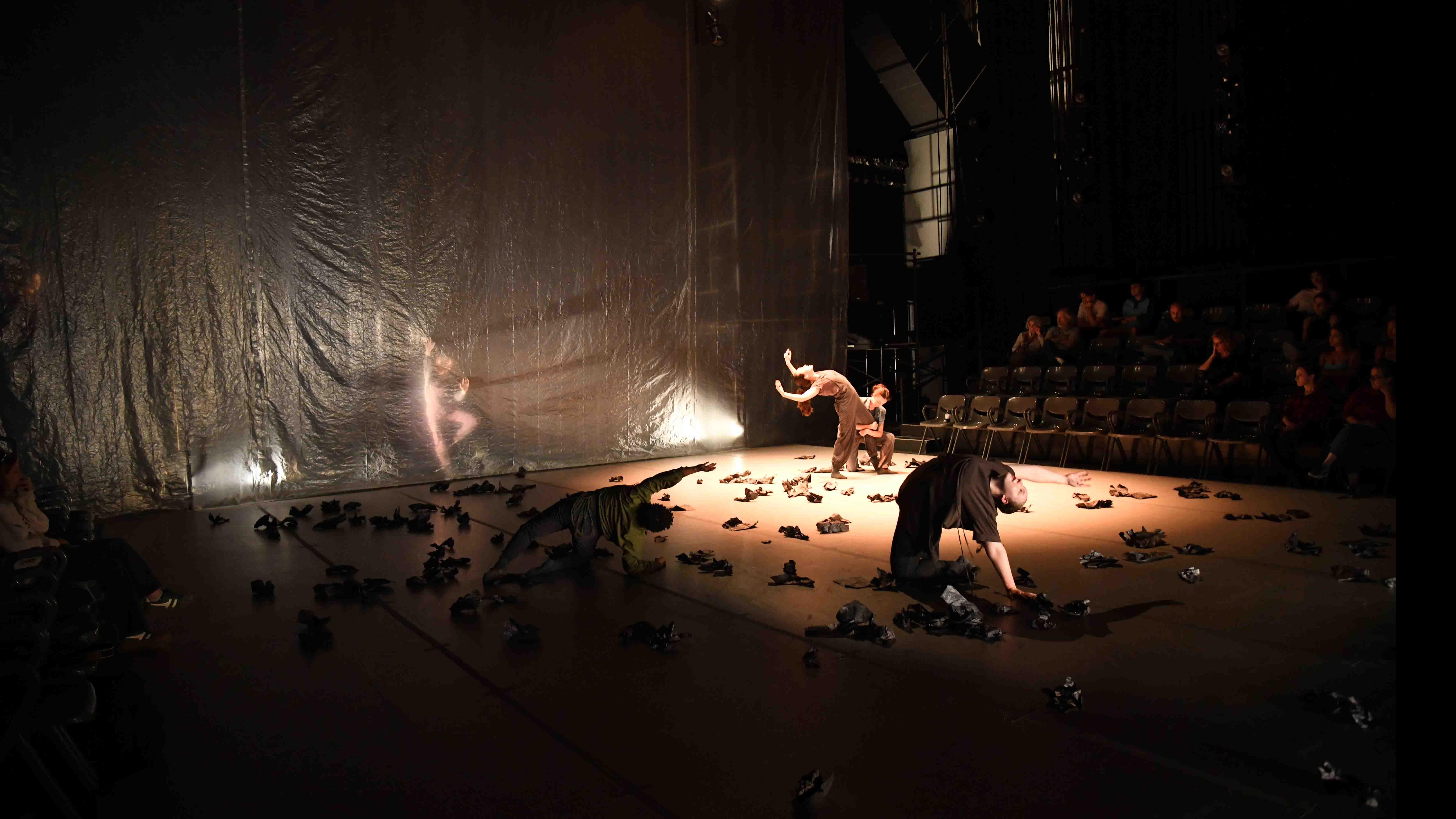 Five student dancers in the foreground and background in various movement poses, with debris on the stage, and a transparent sheet in front of one dancer, with a crowd of people in the back on the right. Photo credit: Thomas Hoebbel