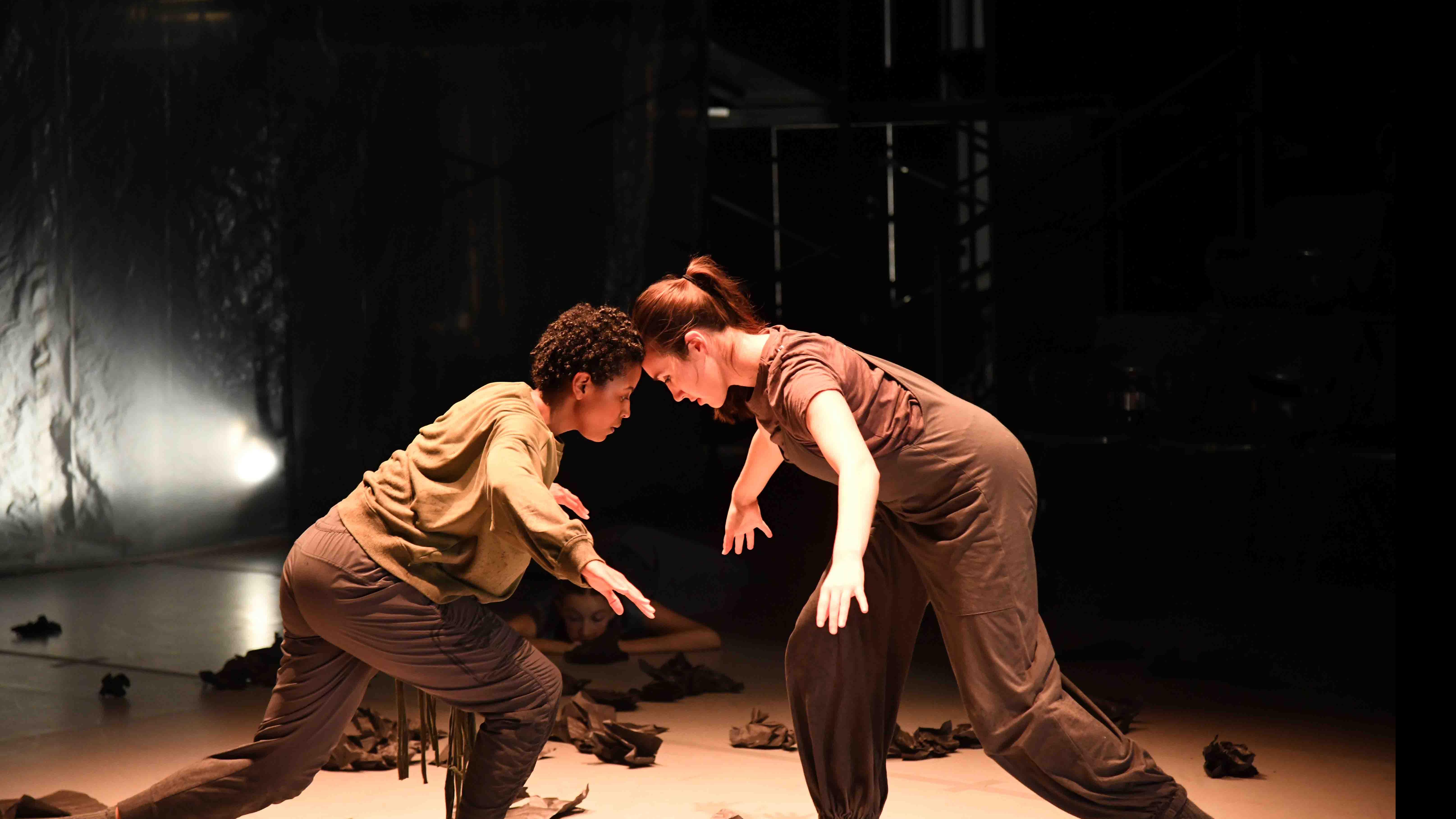 Two student dancers lean forward and press their heads together amidst debris on stage. Photo credit: Thomas Hoebbel