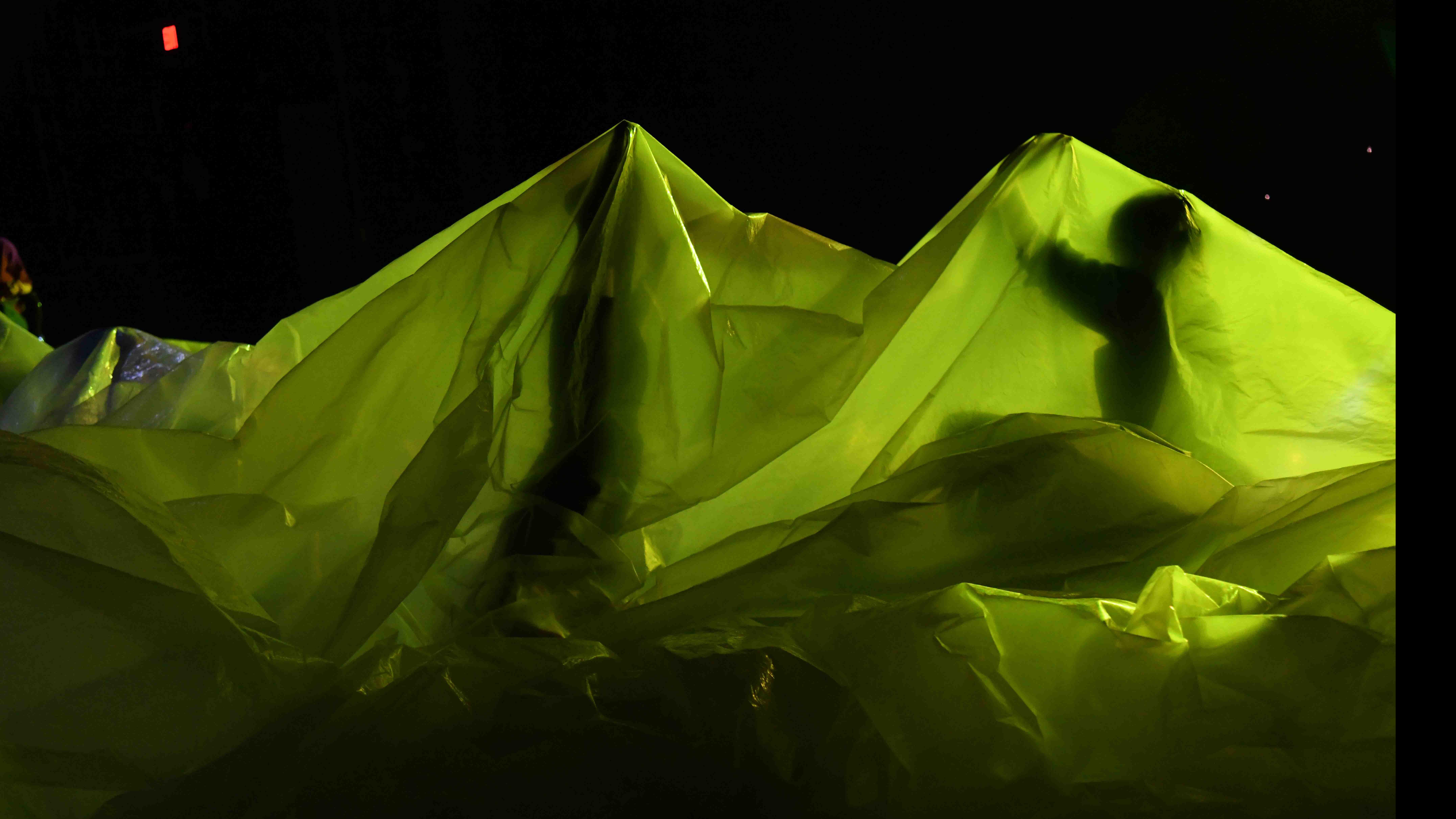 Two student dancers in movement poses beneath a translucent sheet under green light on stage. Photo credit: Thomas Hoebbel