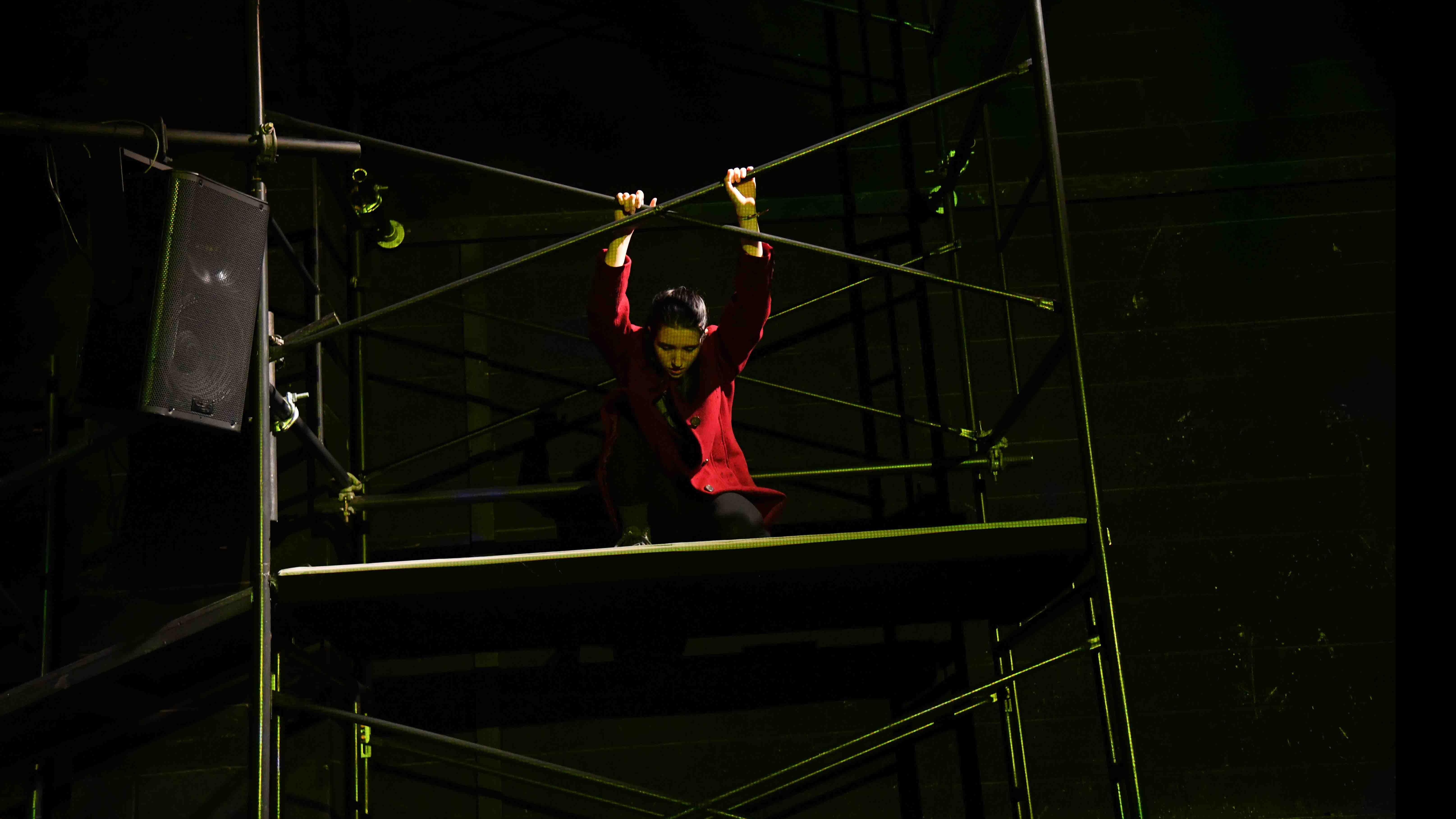 A student dancer in a movement pose holds the bars on a platform on stage. Photo credit: Thomas Hoebbel
