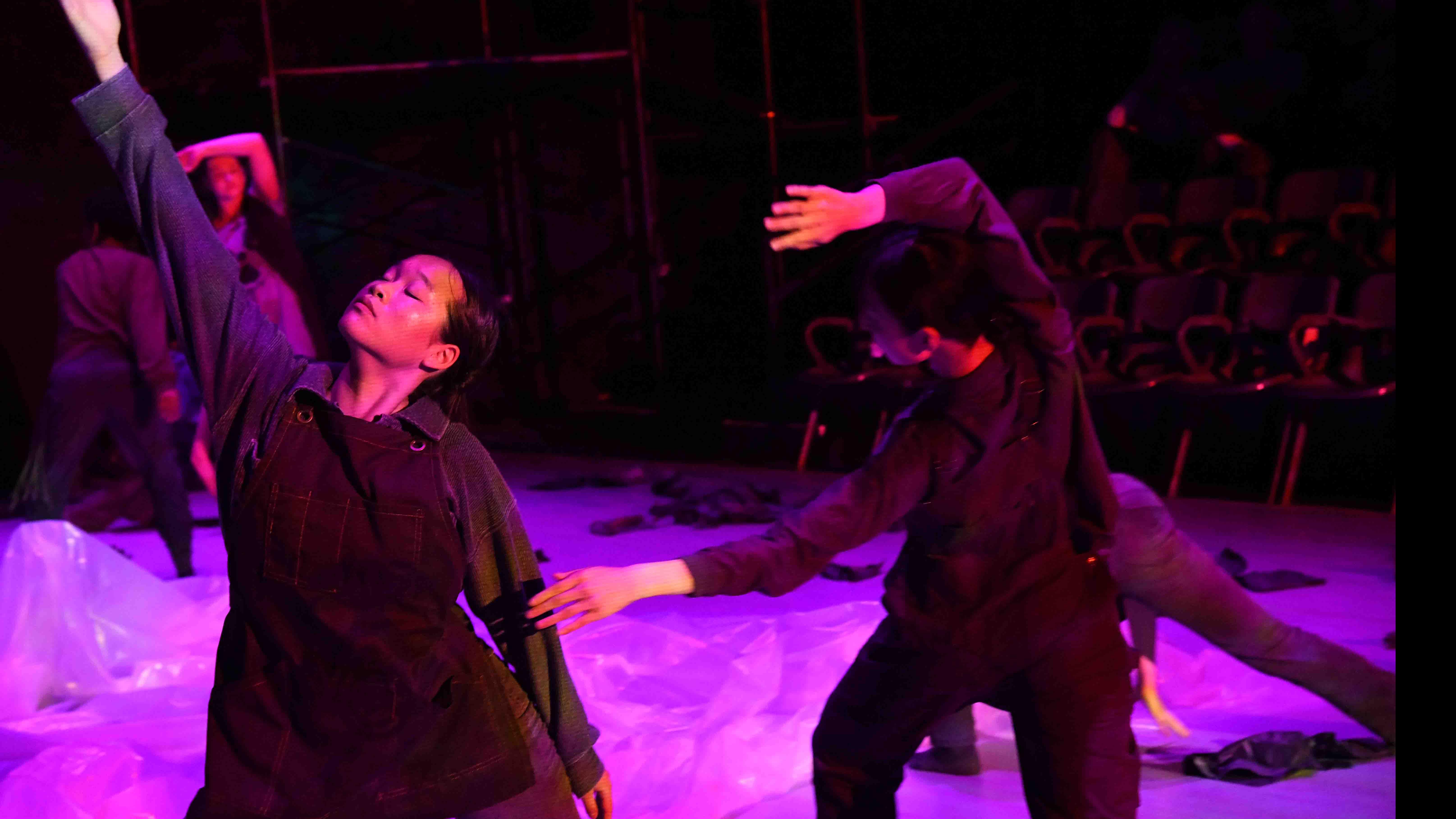 Two student dancers in the foreground in movement poses, and three student dancers in the background in movement poses, with a translucent sheet, debris, and purple light on stage. Photo credit: Thomas Hoebbel