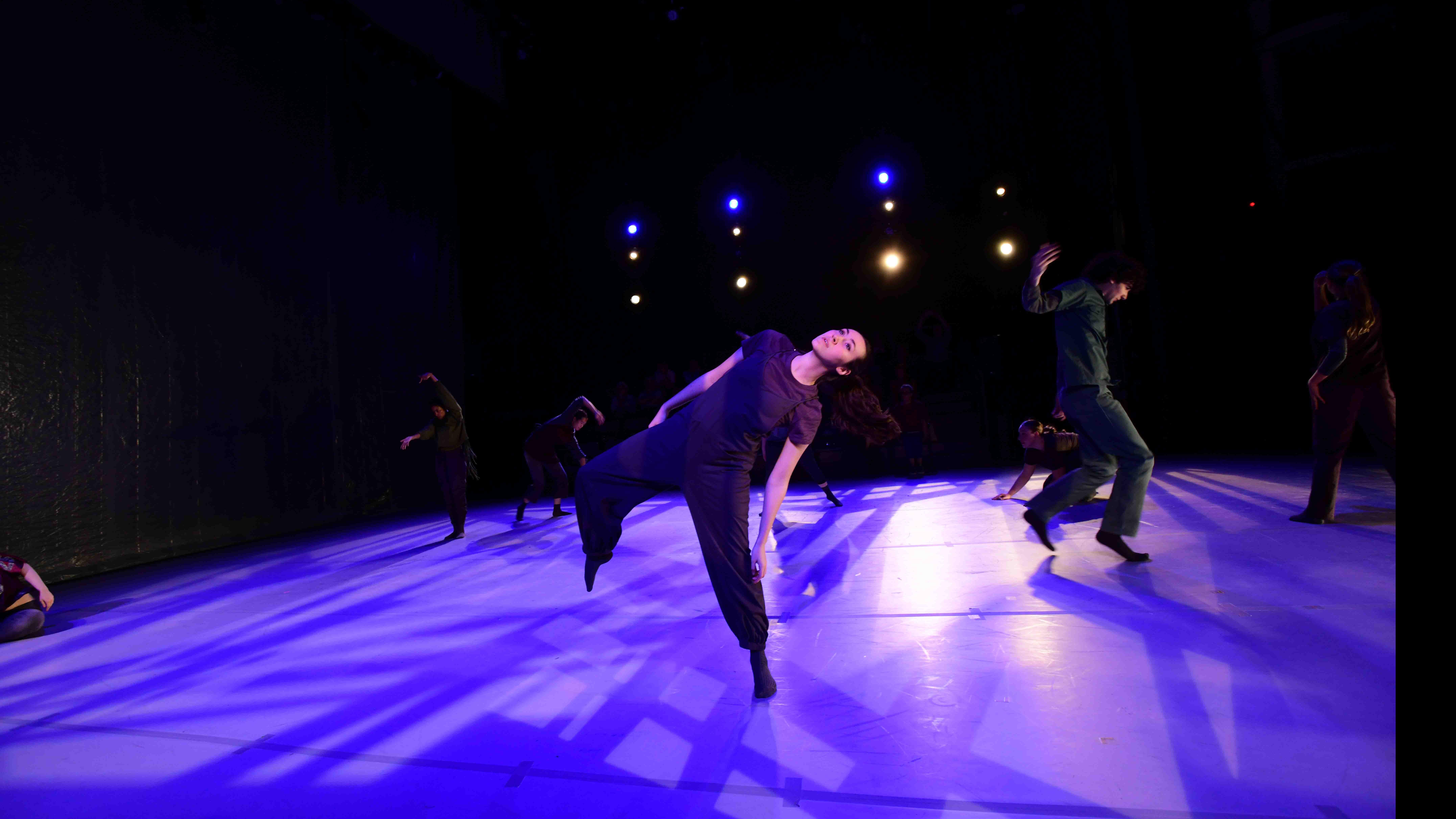An ensemble of student dancers in various movement poses on a stage lit with purple light. Photo credit: Thomas Hoebbel