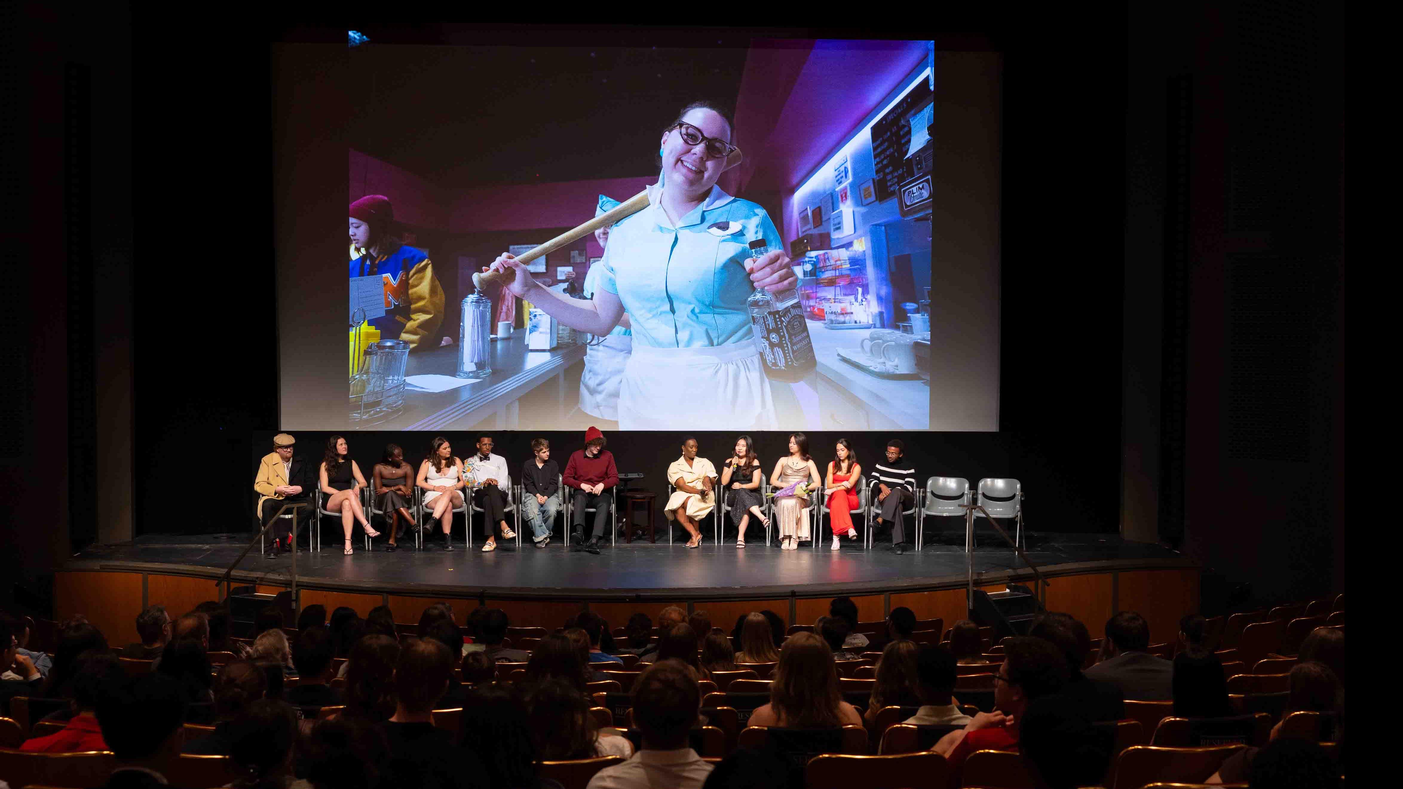 Student cast and crew members of the PMA Studios short film Milkshake Kiss sit on stage in the Kiplinger Theatre, while the audience looks on, and the film plays on a large screen. Photo credit: Chris Kitchen