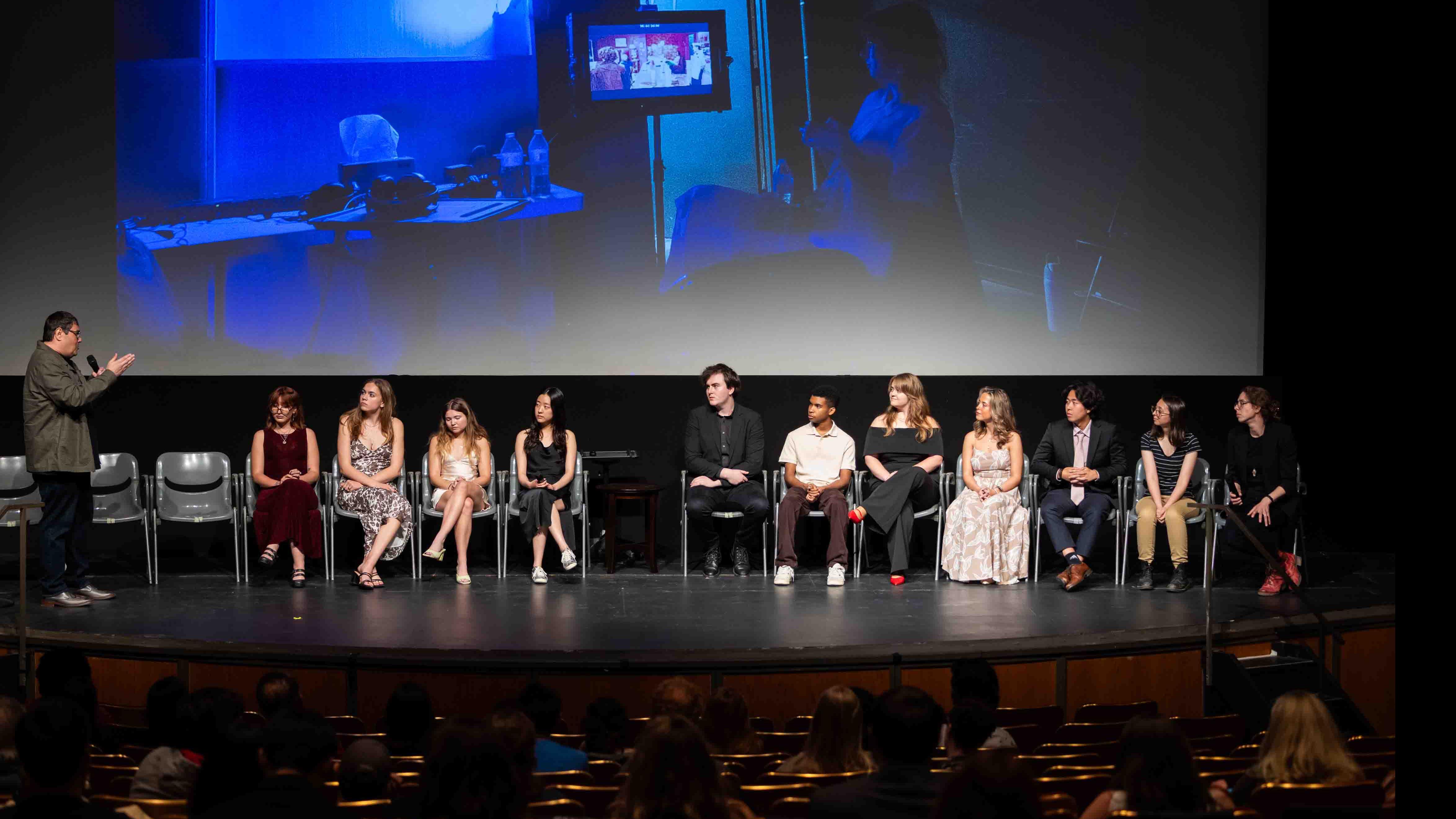 PMA Associate Professor Jeff Palmer speaks into a microphone to a group of student cast and crew members of the PMA Studios short film Milkshake Kiss on stage in the Kiplinger Theatre. Photo credit: Chris Kitchen