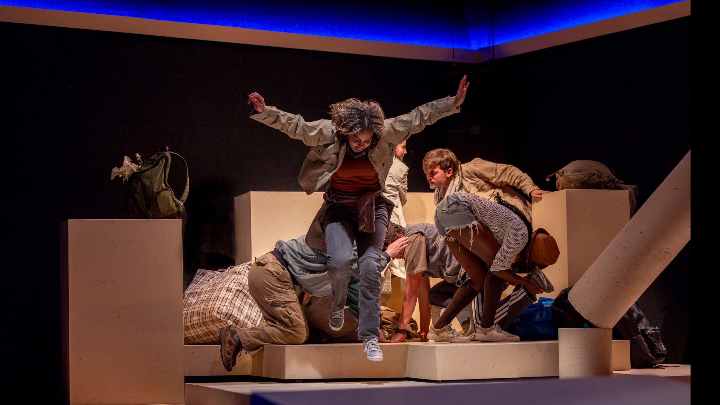 An ensemble of performers huddles together while a cast member jumps off a platform during a performance of I Want a Country in the Flex Theatre, Schwartz Center for the Performing Arts.