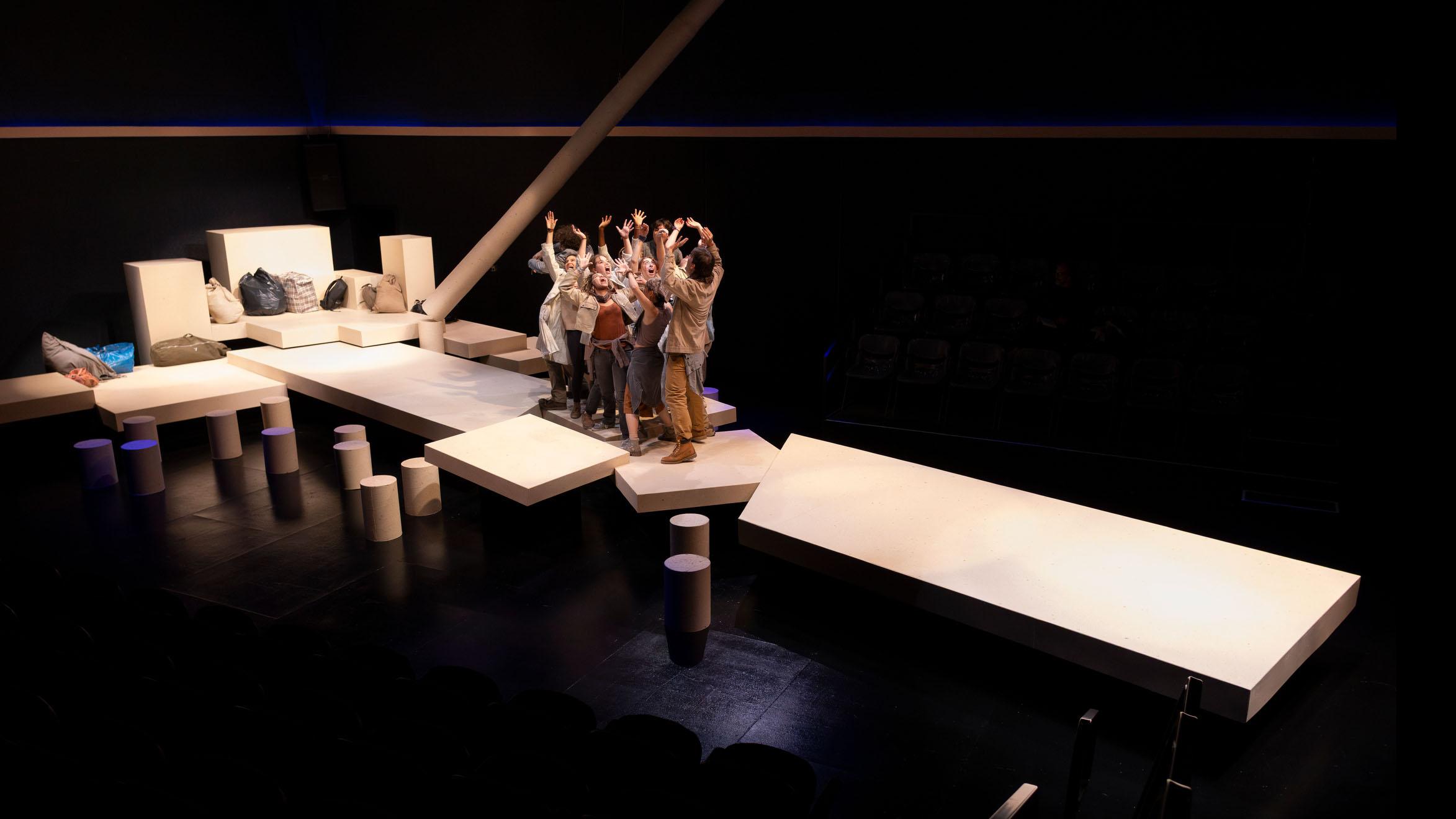 A wide shot of an ensemble of performers grouped tightly together on a long stage made of a series of pieces of wood during a performance of I Want a Country in the Flex Theatre, Schwartz Center for the Performing Arts.