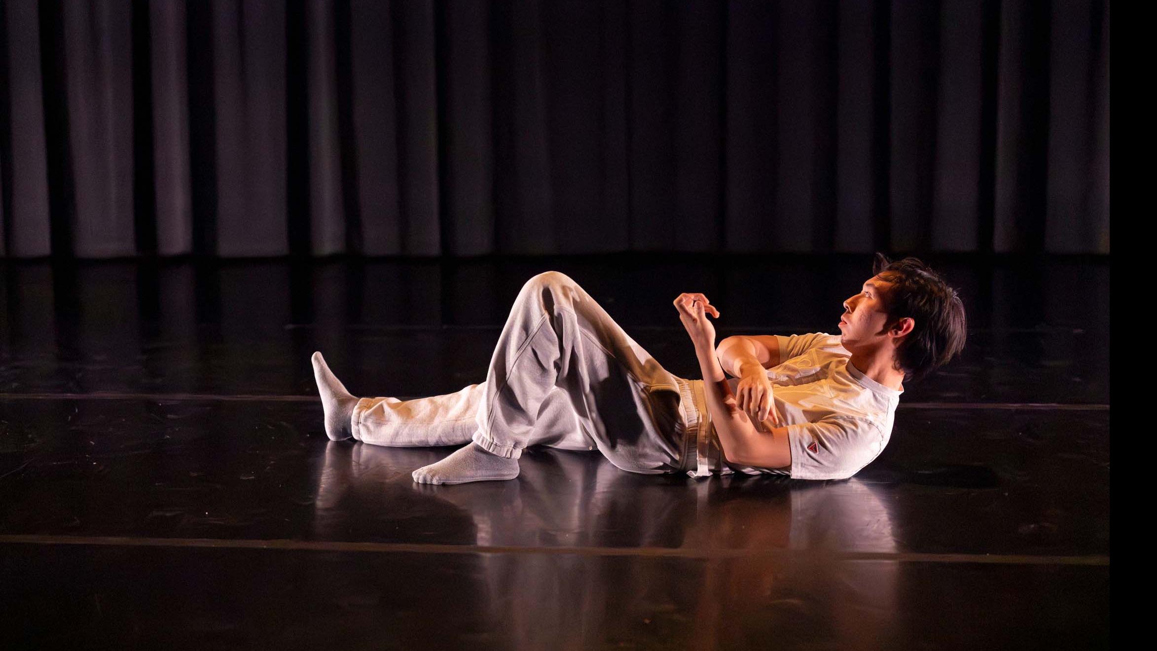 Daniel Si lies on the ground in a movement pose during Share In / Share Out in the Class of ’56 Dance Studio Theatre, Schwartz Center.