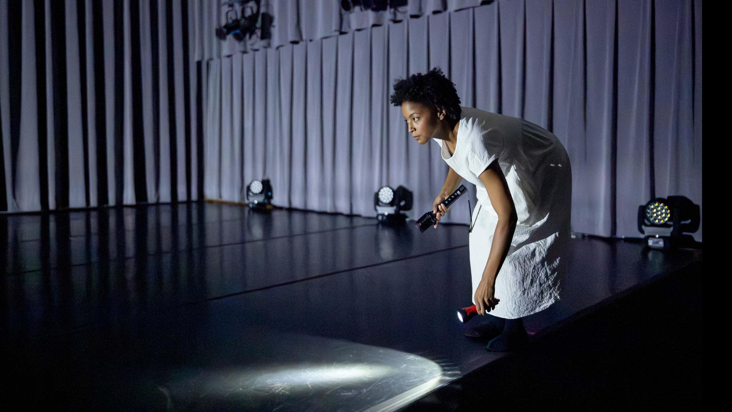 Taylor Janeen Pryor shines a flashlight on the ground in a movement pose during Share In / Share Out in the Class of ’56 Dance Studio Theatre, Schwartz Center.