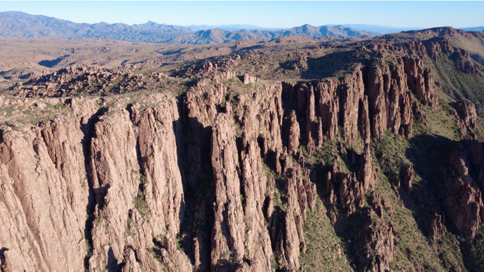 Photograph showing a rugged cliff face with vertical rock formations and sparse vegetation in a desert landscape. Background includes distant mountain ranges under a clear blue sky, highlighting geological features and natural terrain.
