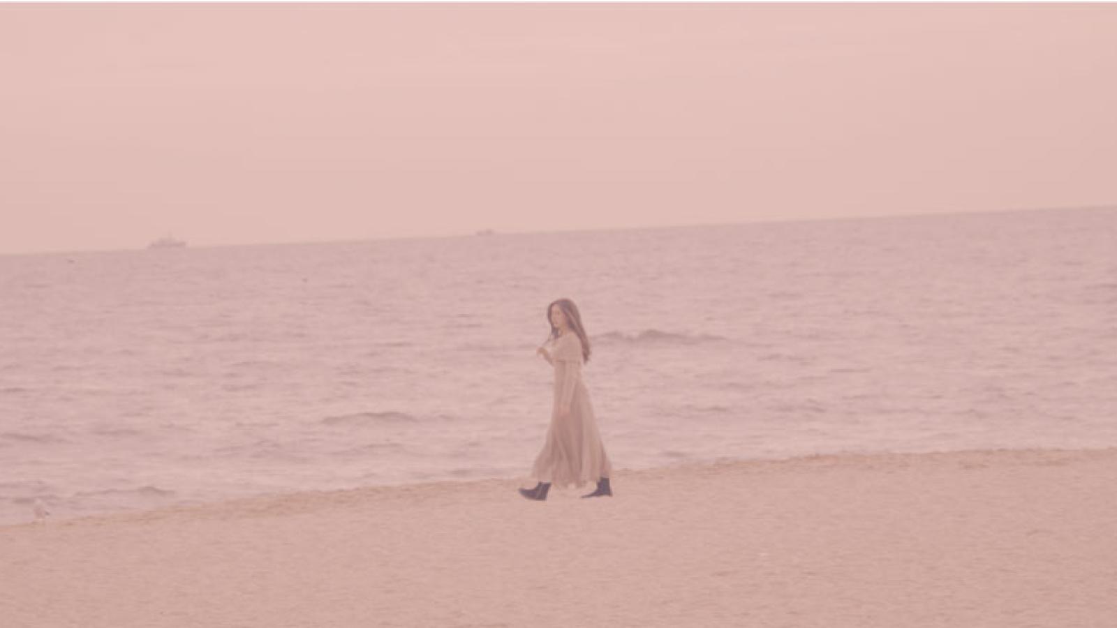 Photograph of a person walking alone on a sandy beach near calm ocean waters under a pale pink sky. The scene conveys a serene and contemplative mood with muted colors and minimal background elements.