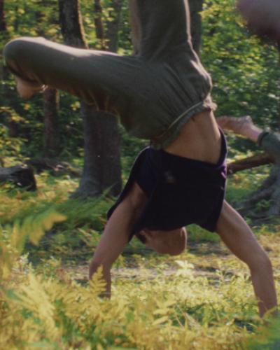 A still from Branché, a short film by Janique L. Robillard and Eric Bates, where a person is carthwheeling through grass with trees in the background