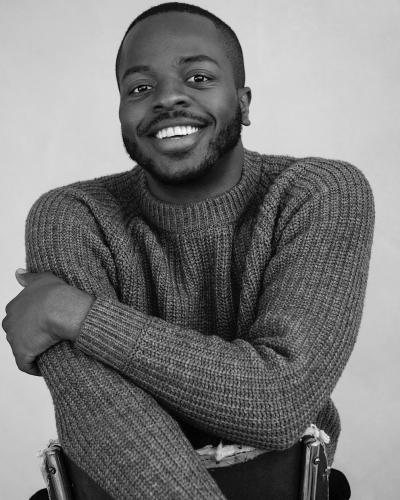 Black-and-white portrait of a Antuan Byers in a chunky knit sweater, seated in a chair turned backward with arms folded over the backrest.