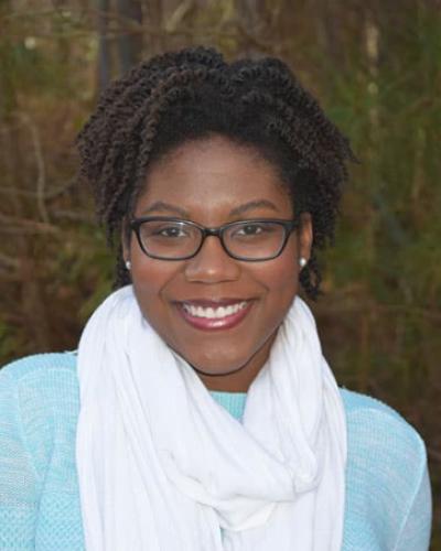 Carley Robinson wearing glasses, a pale blue shirt, and white scarf, with trees visible in the background