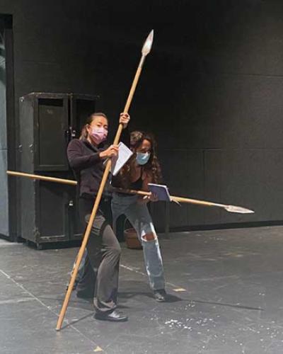 Two actresses holding spears on stage for a Seven Homeless Mammoths Wander New England rehearsal